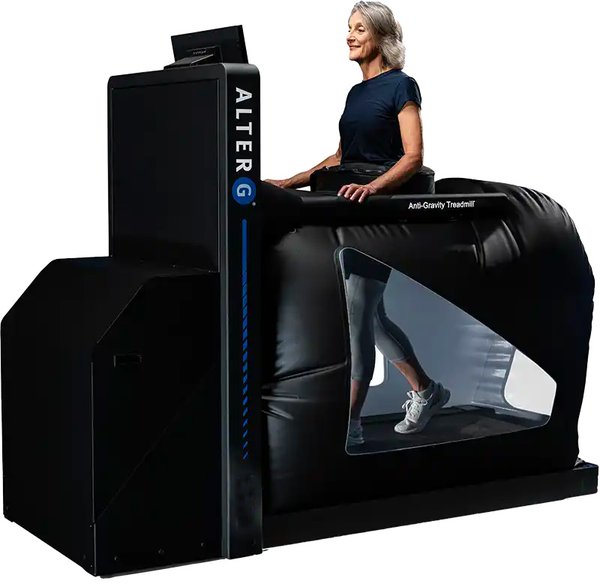 Enclosed treadmill with a pressurized air chamber around the lower body; a woman stands inside, waist-deep, with a clear window showing her legs walking on the belt.